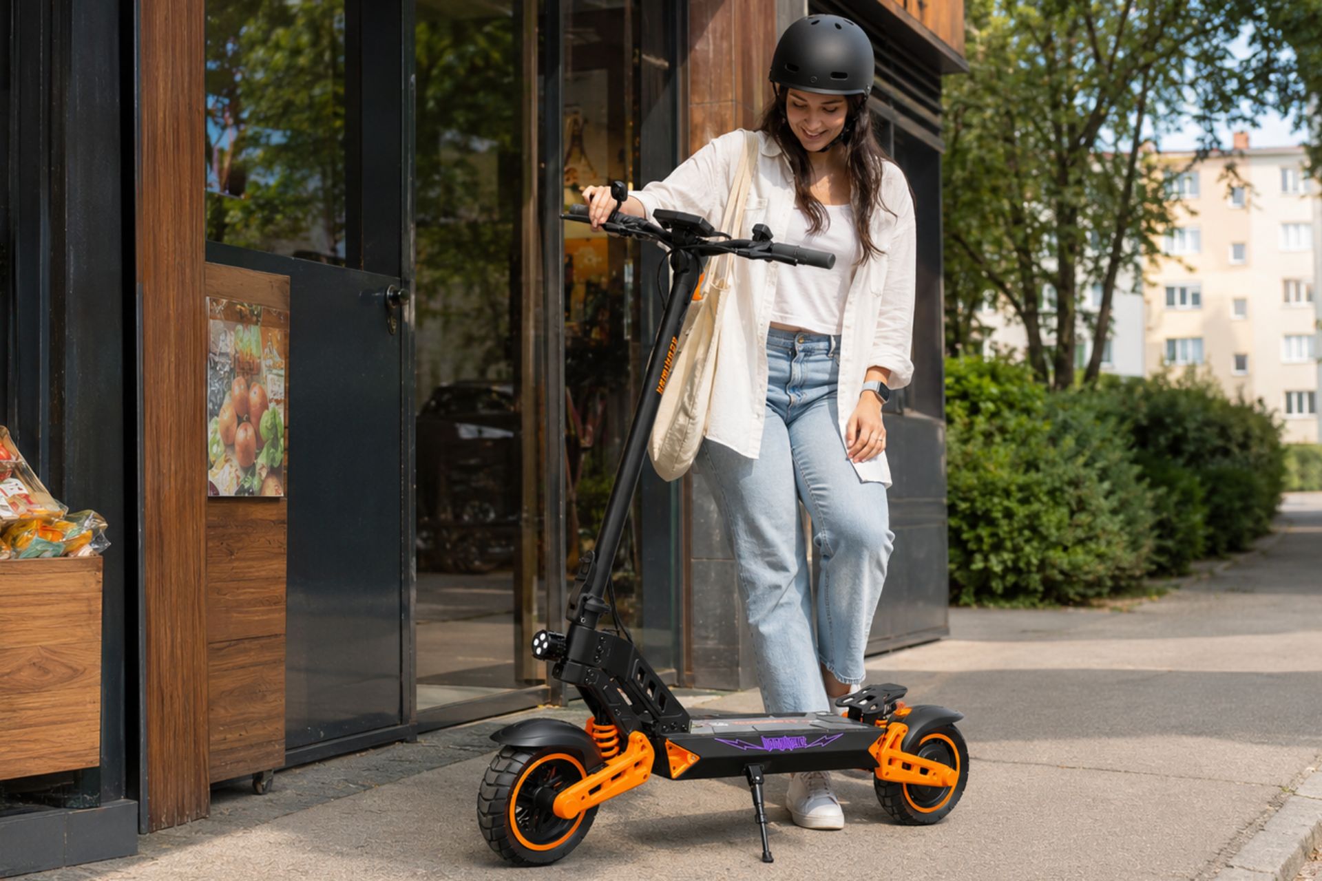kobieta w kasku typu orzeszek na głowie parkuje hulajnogę elektryczną Kamikaze K1 Lite przed sklepem